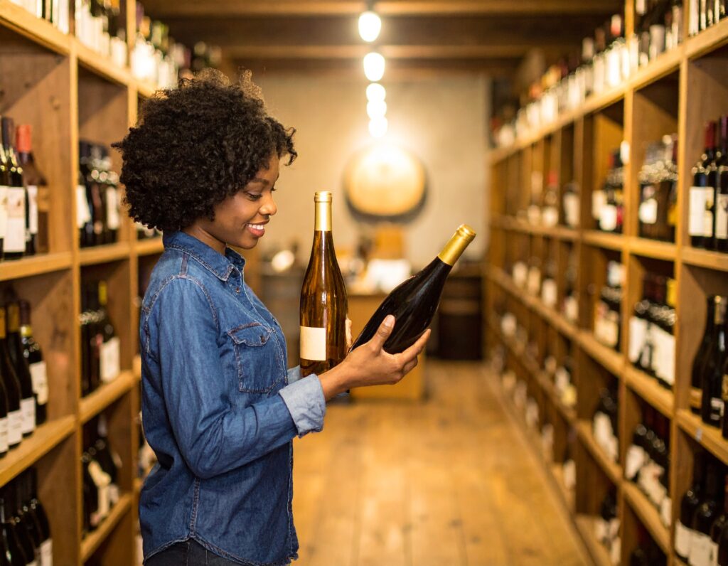 woman standing in an aisle full of wine bottles and holding 2 in her hand and looking at the wine label