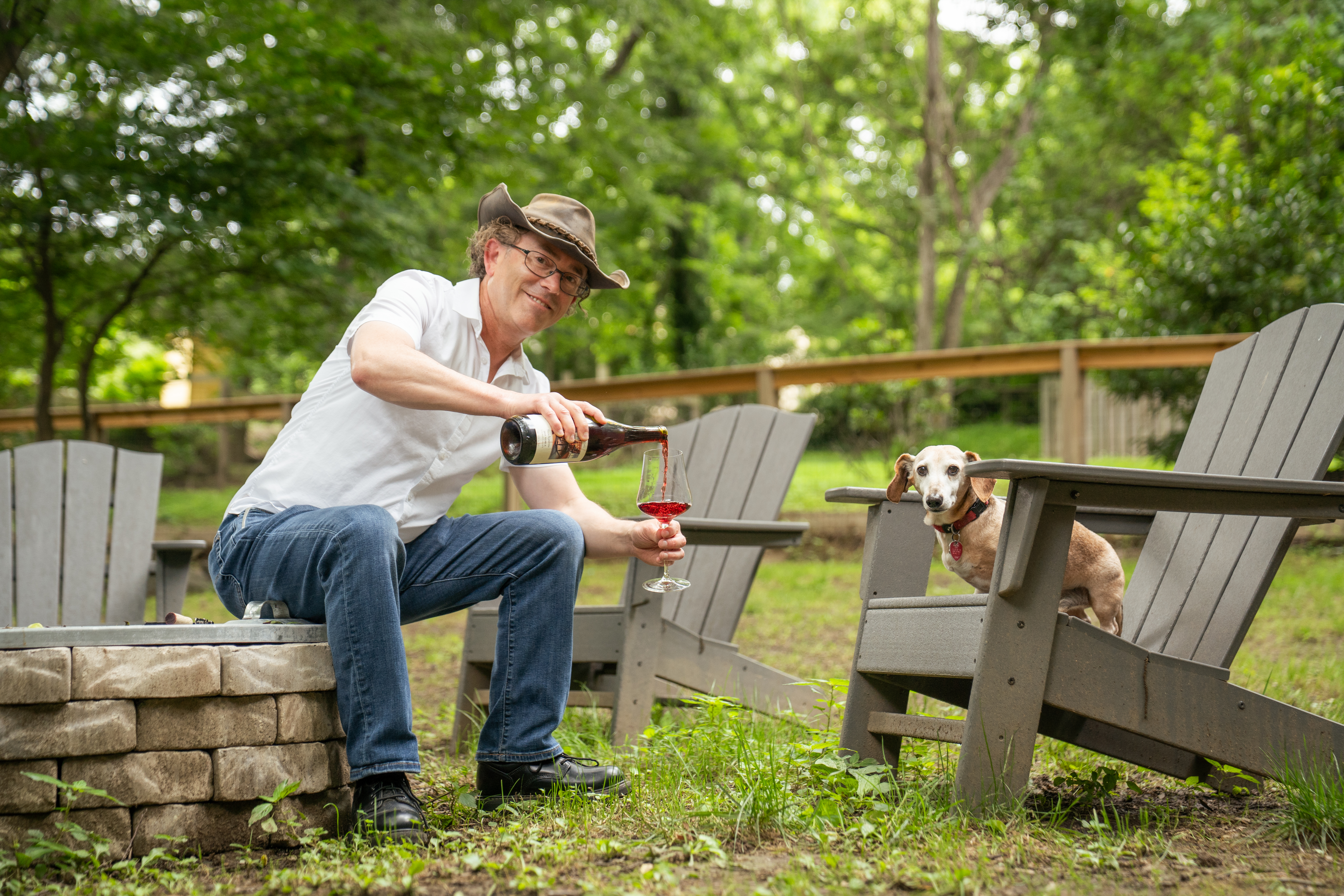 winemaker Eric Martella sitting on a fire pit pouring a glass of wine with his dog watching